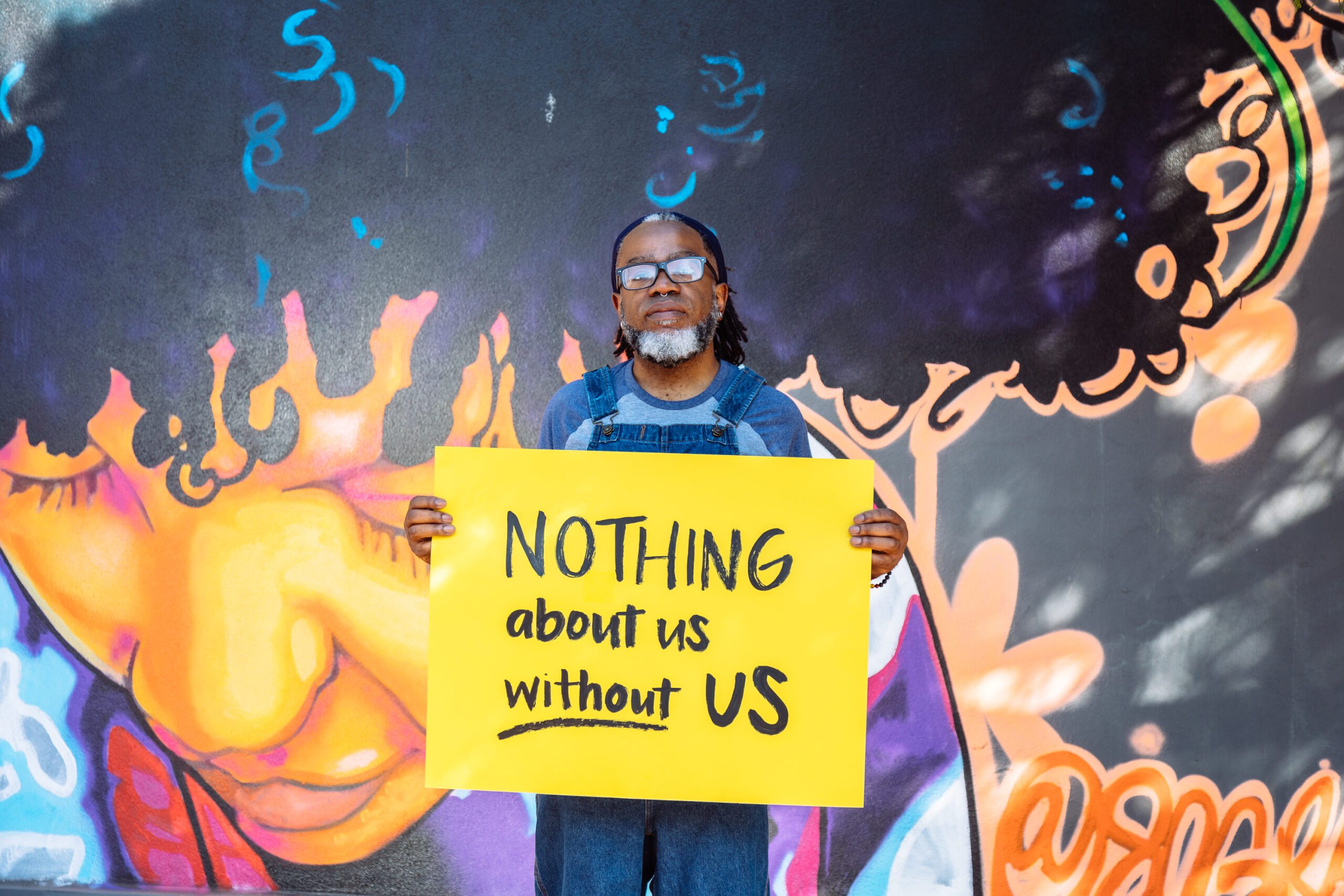 A Deaf Black man wearing glasses looks neutrally at the camera while holding a hand lettered sign declaring “NOTHING about us without US.” The man wears denim overalls and has a black and white beard, septum piercing, and hair pulled back with a bandana. The background features a vibrant mural of a Black woman looking down.