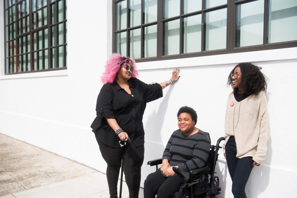 Three Black and disabled folx (a non-binary person resting one hand on the wall and the other on her cane, a non-binary person sitting in a power wheelchair, and a femme leaning while standing) rest and chat outdoors on a sunny day.