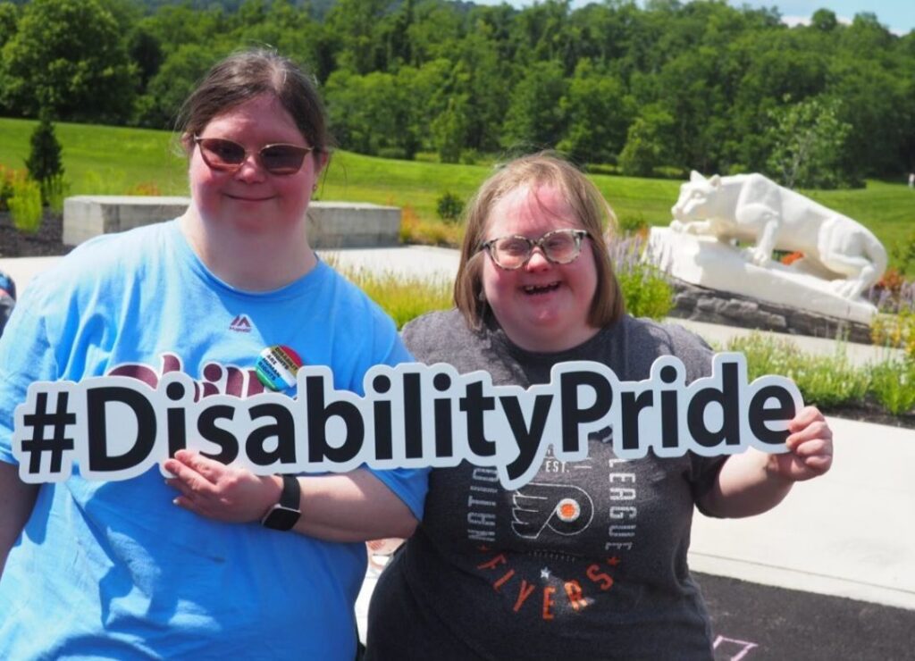 Two women with disabilities standing outside holding a sign that says #DisabilityPride