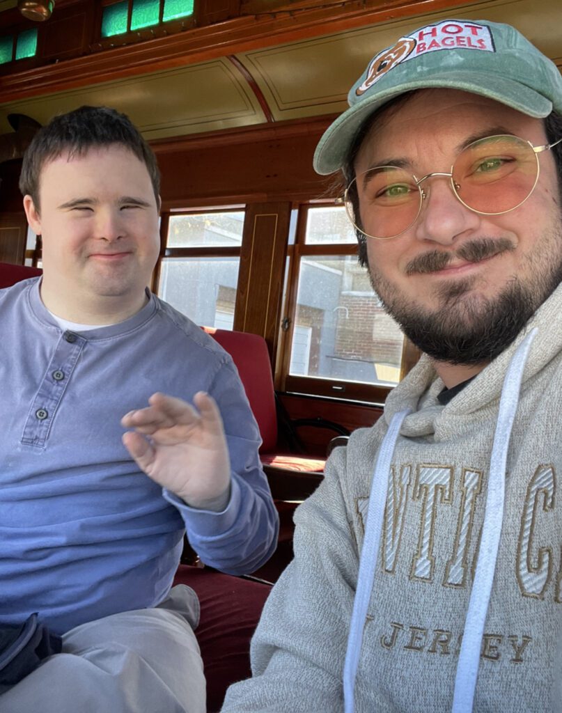 A young man with intellectual disability waves at the camera while in a train with his support partner