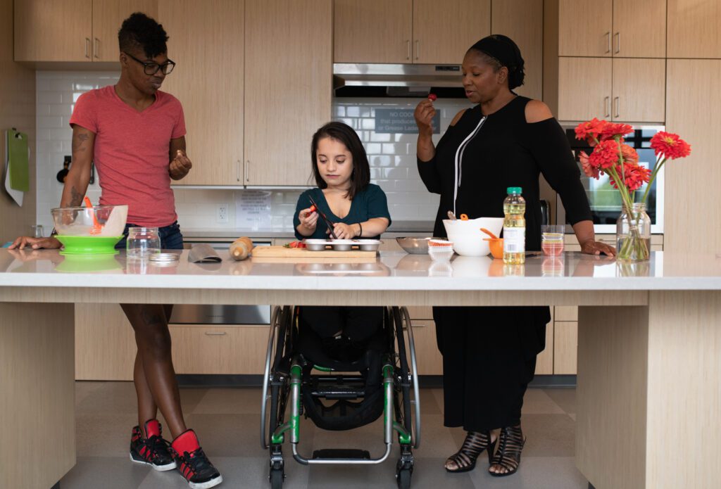 Three disabled people of color (a Black non-binary person on the left, a South Asian person with a wheelchair in the middle, and a Black woman on the right) at a kitchen counter with open space underneath. The person in the middle chops strawberries while the other two each sample a strawberry.