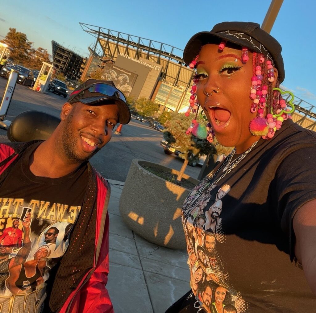 A black man in a wheel chair wearing a baseball cap beside a black woman with bright beads in her hair outside a stadium. 