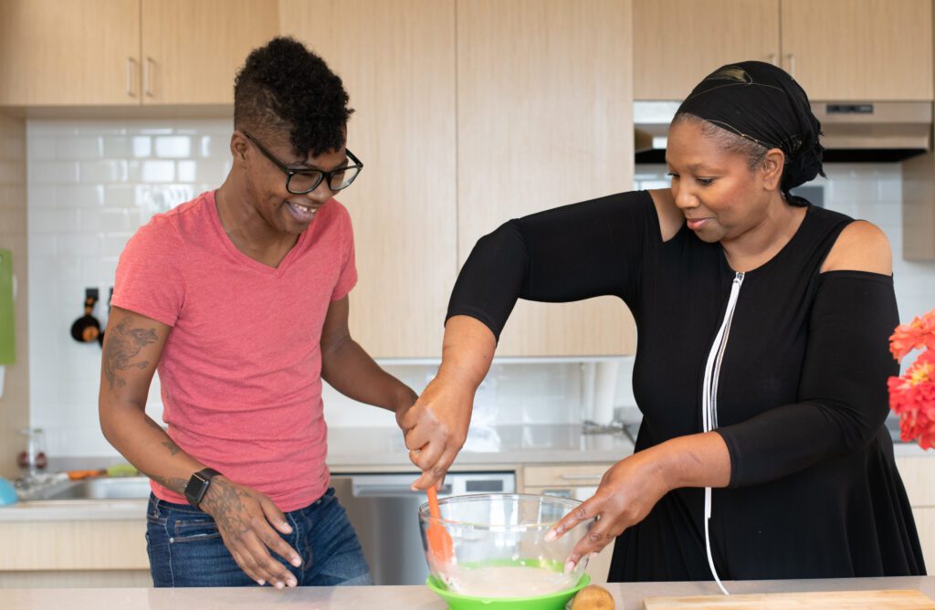 Two disabled and Black people bake together. The woman on the right vigorously stirs a mixing bowl while the non-binary person on the left watches laughingly.