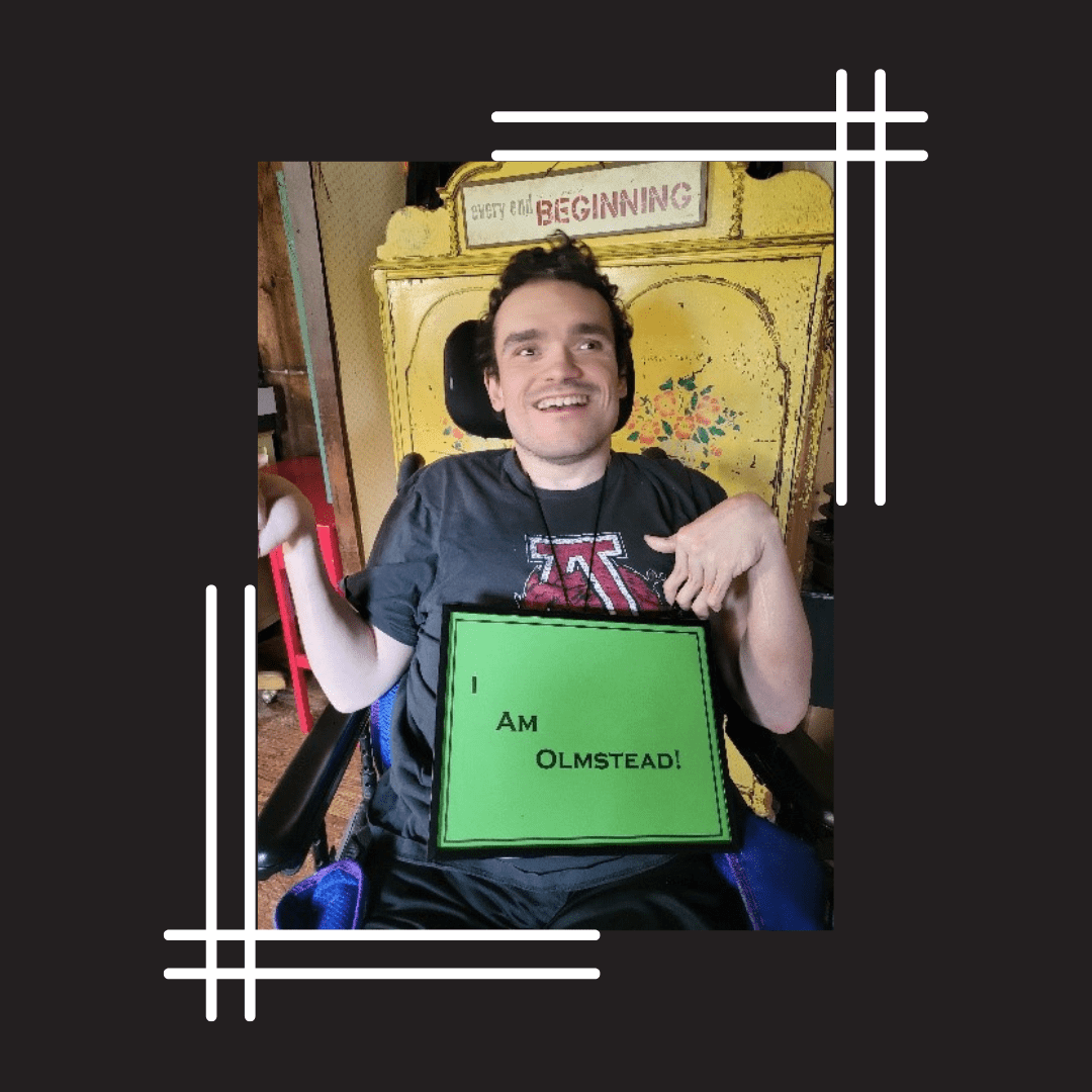 A disabled man smiles brightly at the camera with a green sign saying, "I am Olmstead" on his lap.