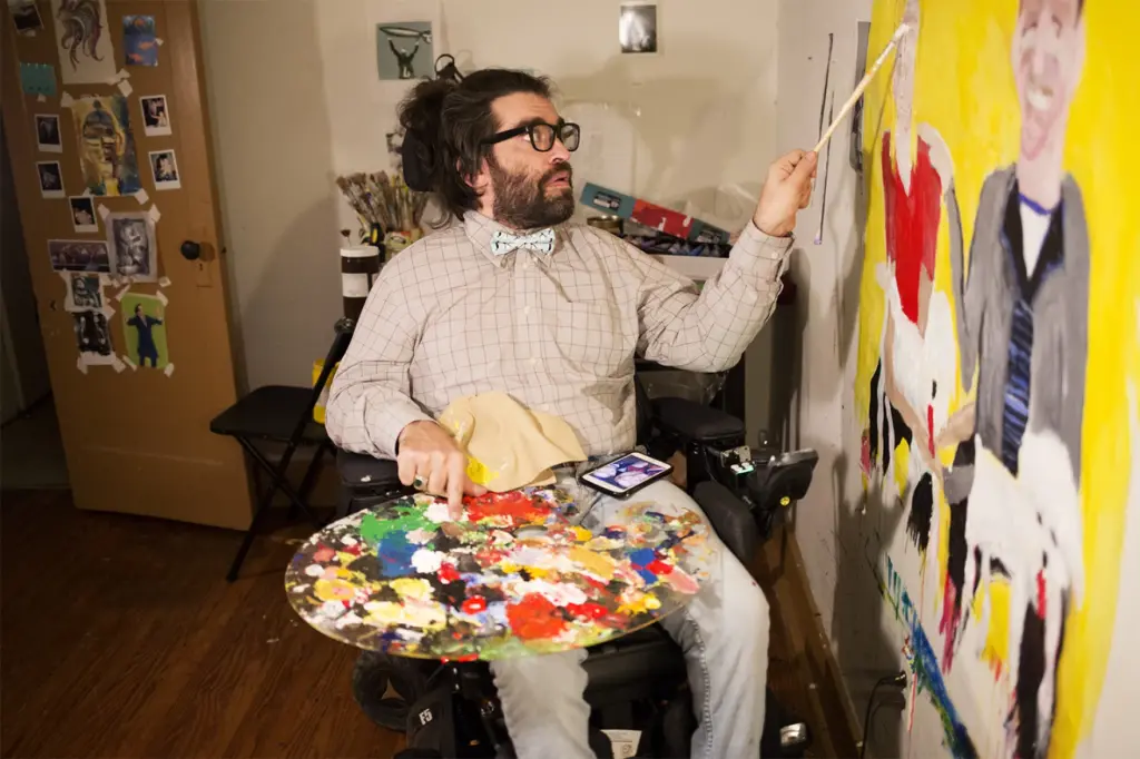 Chris Worth, a disabled white man with a beard and long hair, wears a tan outfit and rests a paint pallet covered in bright colors on his lap as he paints a large picture. 