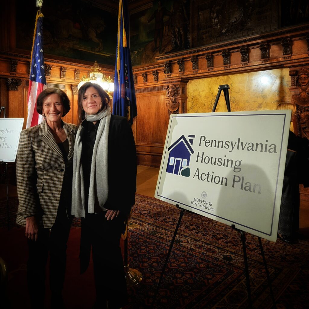 Department of Human Service Secretary Val Arkoosh and Marian Frattarola-Saulino smile at the camera while standing next to a sign that says, "Pennsylvania Housing Action Plan"